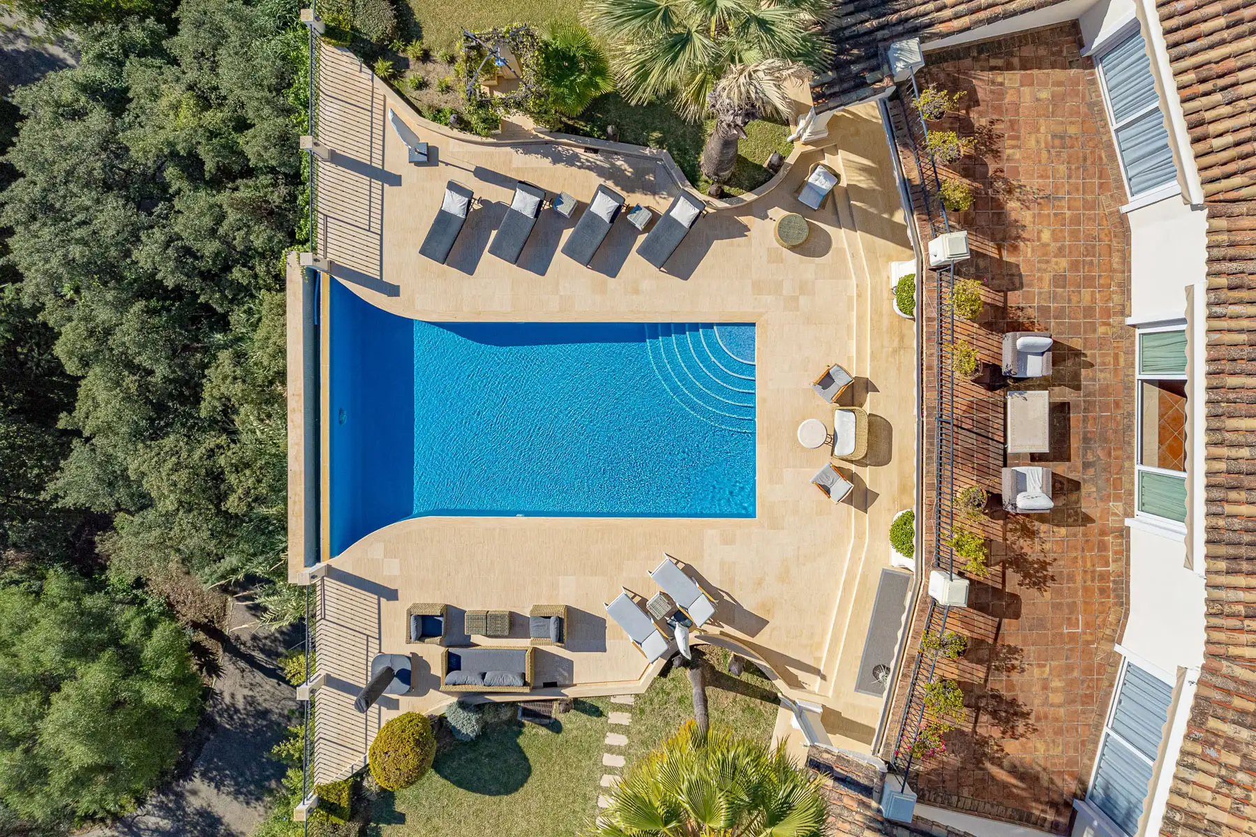 Top-down aerial view of a pool terrace with sun loungers beside a tiled roof and trees