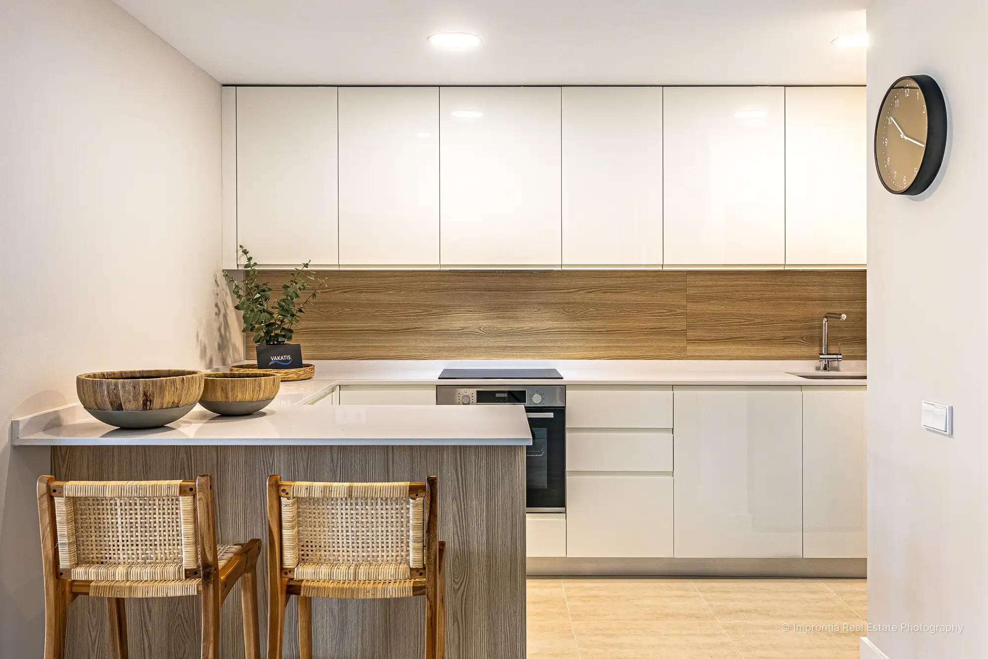 Vista frontal de una cocina con armarios blancos, un frente de madera de roble y dos taburetes de fibra trenzada