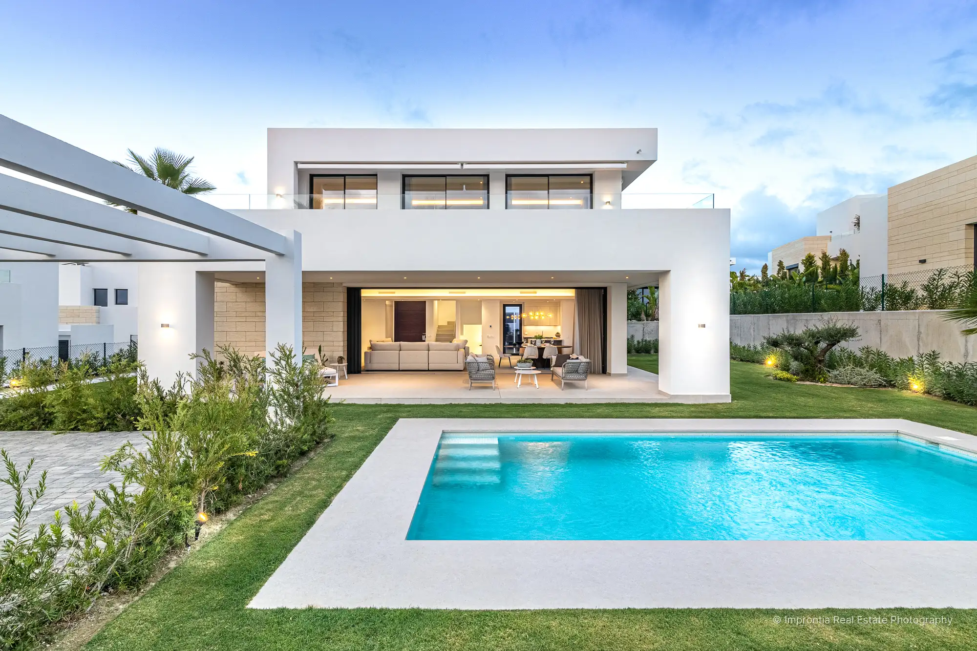 Villa moderna blanca al anochecer con la piscina iluminada, luces cálidas en el interior y una terraza cubierta