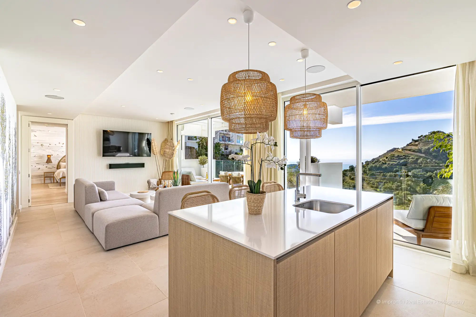Open-plan living room and kitchen with a central island, rattan pendant lamps and a glass wall facing the mountains and the sea