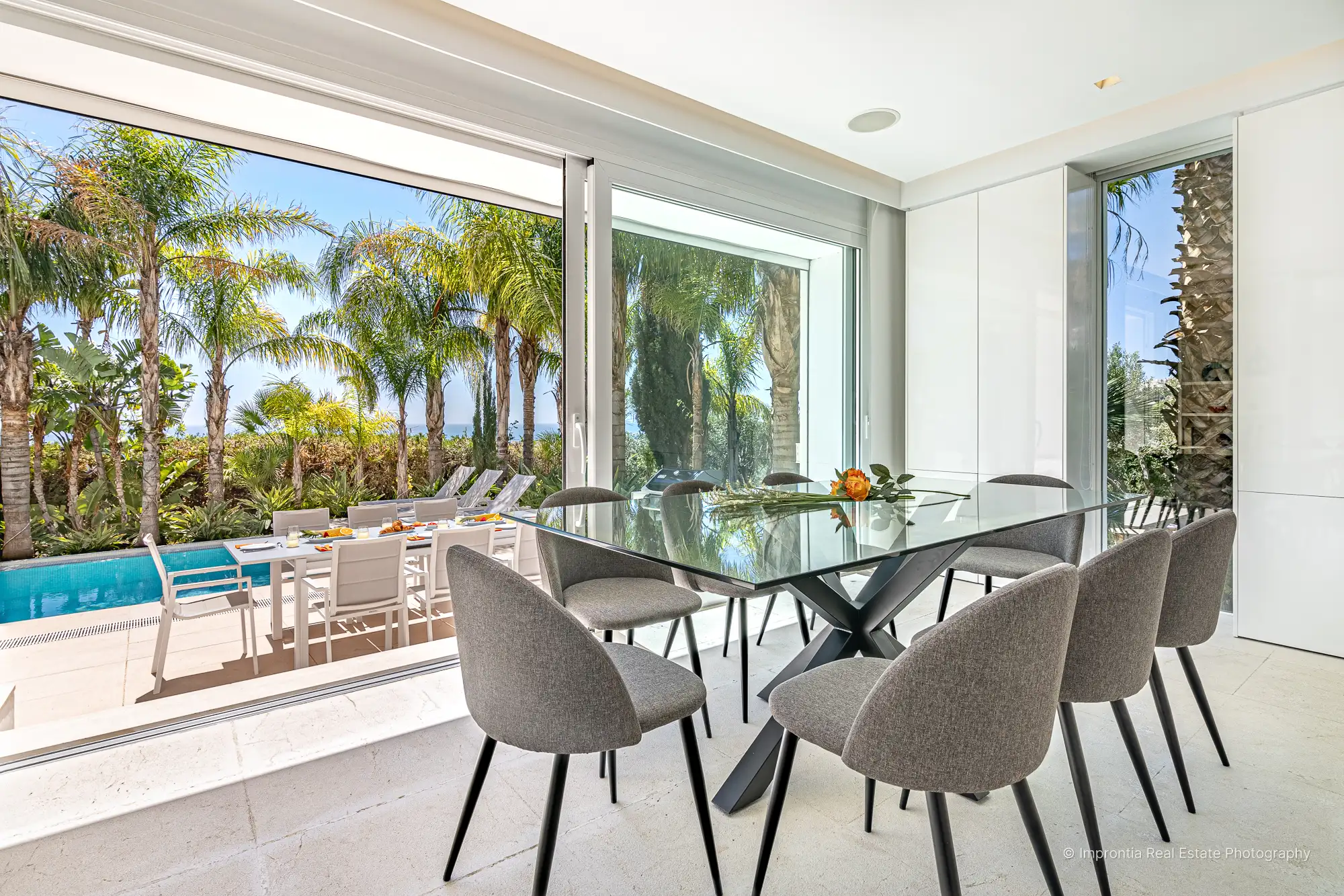 Dining room with a glass table and sliding doors open to a palm garden, a pool and a distant sea view