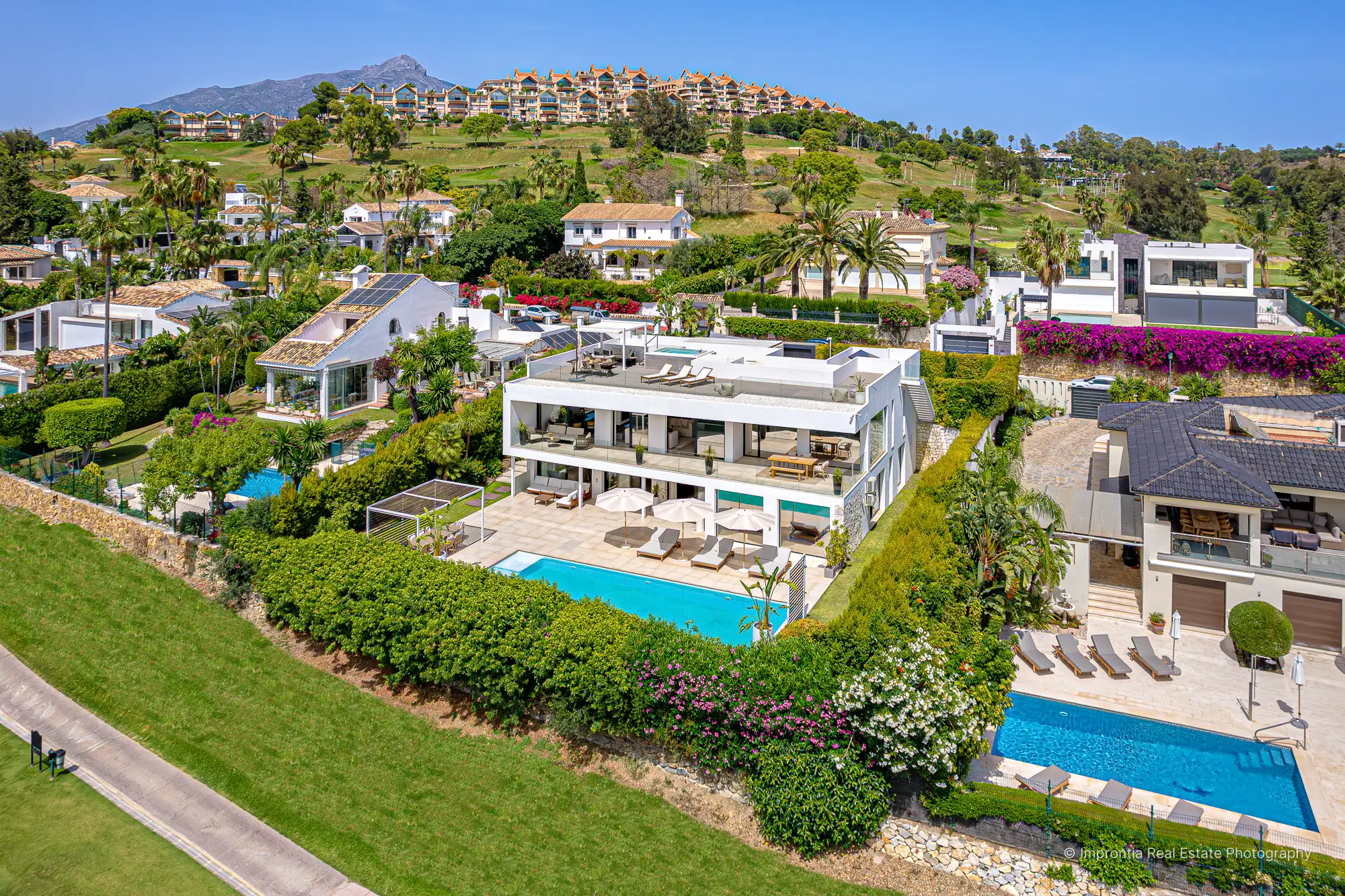 Aerial view of a modern white villa with a rooftop terrace and pool on a golf-course hillside with mountains behind